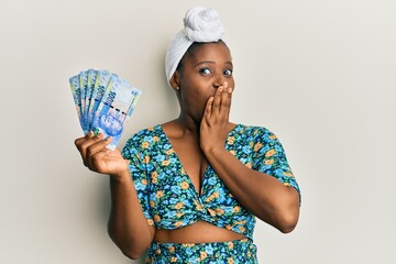 Canvas Print - Young african woman wearing hair turban holding south african 100 rand banknotes covering mouth with hand, shocked and afraid for mistake. surprised expression