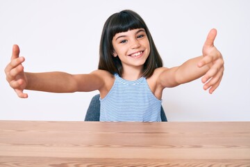 Wall Mural - Young little girl with bang wearing casual clothes sitting on the table looking at the camera smiling with open arms for hug. cheerful expression embracing happiness.