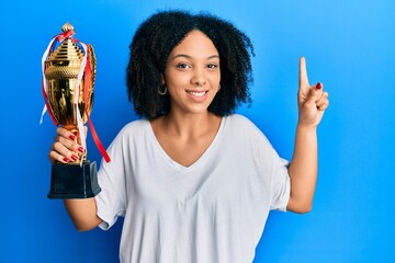 Wall Mural - Young african american girl holding winner trophy smiling with an idea or question pointing finger with happy face, number one
