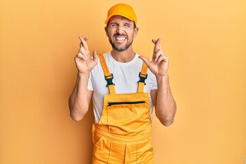 Poster - Young handsome man wearing handyman uniform over yellow background gesturing finger crossed smiling with hope and eyes closed. luck and superstitious concept.