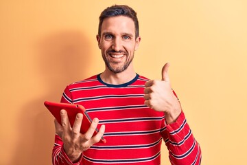 Wall Mural - Young handsome man holding touchpad standing over isolated yellow background smiling happy and positive, thumb up doing excellent and approval sign