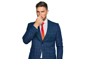 Wall Mural - Handsome man with beard wearing business suit and tie smelling something stinky and disgusting, intolerable smell, holding breath with fingers on nose. bad smell