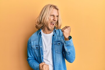Caucasian man with blond long hair wearing casual denim jacket celebrating surprised and amazed for success with arms raised and eyes closed