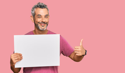 Middle age grey-haired man holding blank empty banner smiling happy and positive, thumb up doing excellent and approval sign