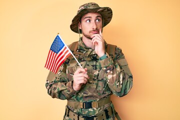 Canvas Print - Young caucasian man wearing camouflage army uniform holding usa flag serious face thinking about question with hand on chin, thoughtful about confusing idea