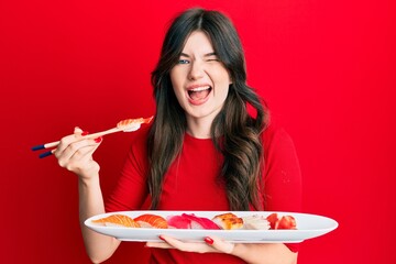 Sticker - Young beautiful caucasian girl eating sushi using chopsticks winking looking at the camera with sexy expression, cheerful and happy face.