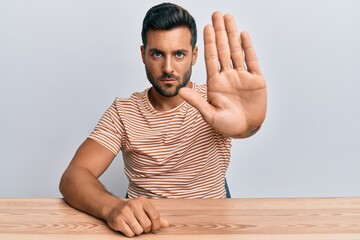 Wall Mural - Handsome hispanic man wearing casual clothes sitting on the table doing stop sing with palm of the hand. warning expression with negative and serious gesture on the face.