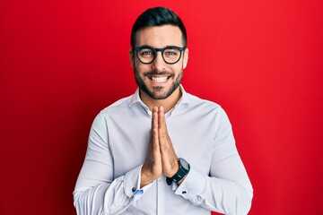 Canvas Print - Young hispanic businessman wearing shirt and glasses praying with hands together asking for forgiveness smiling confident.