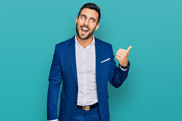 Young hispanic man wearing business jacket smiling with happy face looking and pointing to the side with thumb up.