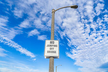 No Jumping Off Bridge Sign On Lamp Post Against Blue Sky And Puffy Clouds