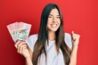 © Krakenimages.com - Young brunette woman holding 100 new zealand dollars banknotes screaming proud, celebrating victory and success very excited with raised arm