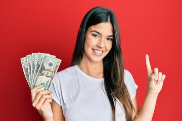 Poster - Young brunette woman holding 20 dollars banknote smiling with an idea or question pointing finger with happy face, number one