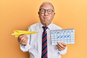 Canvas Print - Senior caucasian man going on business trip holding paper airplane puffing cheeks with funny face. mouth inflated with air, catching air.