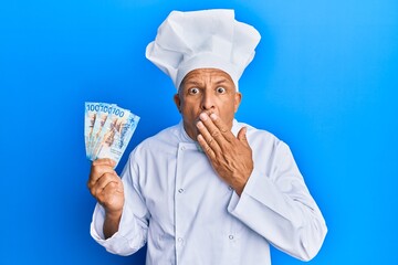 Poster - Middle age grey-haired man wearing professional cook uniform holding swiss franc banknotes covering mouth with hand, shocked and afraid for mistake. surprised expression