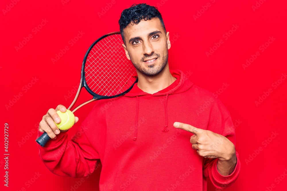 Young hispanic man playing tennis holding racket and ball smiling happy pointing with hand and finger