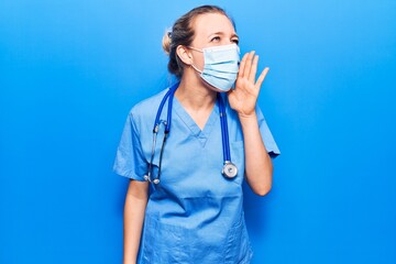 Wall Mural - Young blonde woman wearing doctor uniform and medical mask shouting and screaming loud to side with hand on mouth. communication concept.