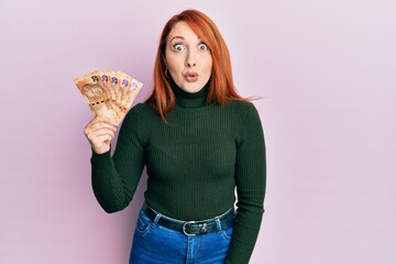 Poster - Beautiful redhead woman holding south african 20 rand banknotes scared and amazed with open mouth for surprise, disbelief face