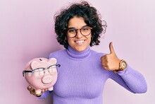 Young Hispanic Woman With Curly Hair Holding Piggy Bank With Glasses Smiling Happy And Positive, Thumb Up Doing Excellent And Approval Sign