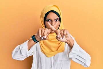 Poster - Young brunette arab woman wearing traditional islamic hijab scarf rejection expression crossing fingers doing negative sign