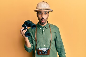 Wall Mural - Young hispanic man wearing explorer hat looking through binoculars scared and amazed with open mouth for surprise, disbelief face