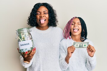 Wall Mural - Beautiful african american mother and daughter holding jar with savings and 1 dollar banknote sticking tongue out happy with funny expression.