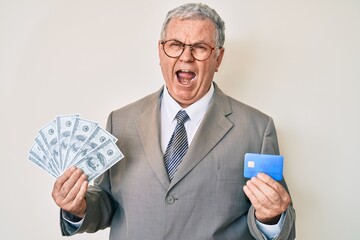 Canvas Print - Senior grey-haired man wearing business suit holding credit car and dollars angry and mad screaming frustrated and furious, shouting with anger. rage and aggressive concept.