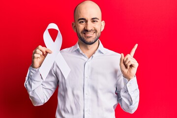 Sticker - Young handsome bald man holding white cancer ribbon smiling happy pointing with hand and finger to the side