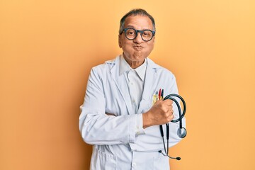 Wall Mural - Middle age indian man wearing doctor coat holding stethoscope puffing cheeks with funny face. mouth inflated with air, catching air.