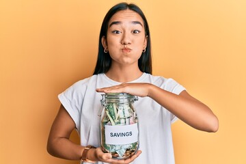 Sticker - Young asian woman holding jar with savings puffing cheeks with funny face. mouth inflated with air, catching air.