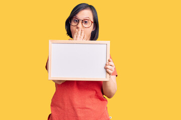 Poster - Brunette woman with down syndrome holding empty white chalkboard covering mouth with hand, shocked and afraid for mistake. surprised expression