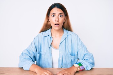 Wall Mural - Young brunette woman sitting on the table wearing casual clothes scared and amazed with open mouth for surprise, disbelief face