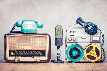 Poster - Retro broadcast radio, mint blue telephone, old microphone, outdated reel to reel tape recorder circa 60s and headphones on wooden table front concrete wall background. Vintage style filtered photo