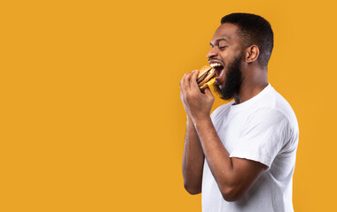 Wall Mural - Black Millennial Guy Biting Burger Eating Over Yellow Background, Side-View