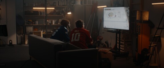 Two friends playing hockey sport video game inside garage hideout, enjoying pizza and drinks. TV screen is blurred