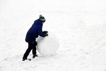 Child Rolls A Snowball On A Street. Winter Leisure, Boy Making A Snowman