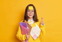 Pleased Asain Female Student Believes In Good Luck At Exam Stands With Eyes Closed And Fingers Crossed Believes Dreams Come True Stuck With Papers Holds Folders Isolated Over Yellow Background