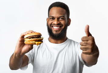 Wall Mural - Black Man Holding Burger And Gesturing Thumbs-Up Over Yellow Background