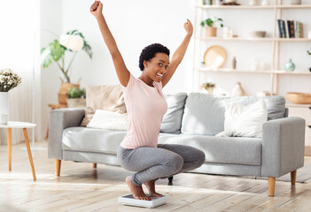 Young black woman sitting on scales, raising hands up in excitement, happy with result of her weight loss diet at home