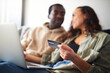 © Southworks - Young Couple At Home Sitting On Sofa Holding Credit Card Using Laptop To Shop Online