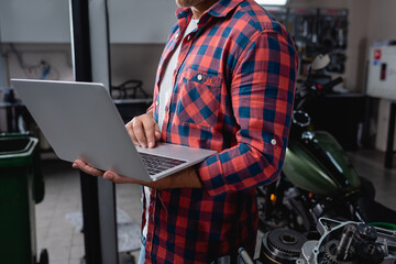 cropped view of technician in plaid shirt with laptop near motorcycle spare parts in garage