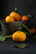 © matyuschenko - Several ripe tangerines with leaves in a metal container lie on a black table against a dark background. Rustic style with copyspace.