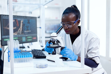 Wall Mural - African researcher adjusting microscope lenses looking at sample on glass slide. Black healthcare scientist in biochemistry laboratory wearing sterile equipment.