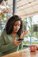 dissatisfied african american woman looking at smartphone while gesturing in cafe