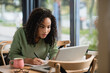 © LIGHTFIELD STUDIOS - african american woman writing in notebook while studying online in cafe