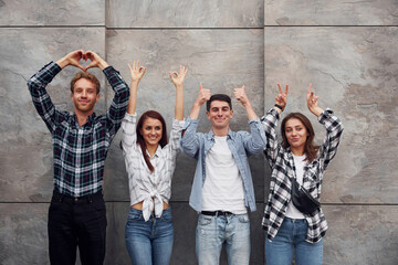 Wall Mural - Making different gestures. Group of young positive friends in casual clothes standing together against grey wall