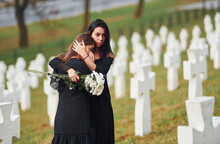 Embracing Each Other And Crying. Two Young Women In Black Clothes Visiting Cemetery With Many White Crosses. Conception Of Funeral And Death