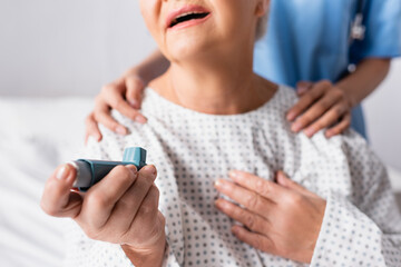 Wall Mural - cropped view of elderly woman holding inhaler while suffering from asthma attack near nurse touching her shoulders, blurred background