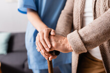 Partial View Of Nurse Touching Hands Of Senior Woman Standing With Walking Stick, Blurred Background