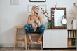 © diignat - Young pretty blonde woman posing at home while relaxing on chair near a white wall having a frame with copy space on background. Modern minimal decor