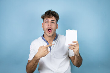 Young handsome man wearing a white t-shirt over blue background smiling and showing blank notebook